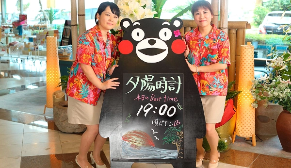 アロハシャツを着た二人の女性が、くまモンの形をした夕陽時刻の看板を挟んで立っています。