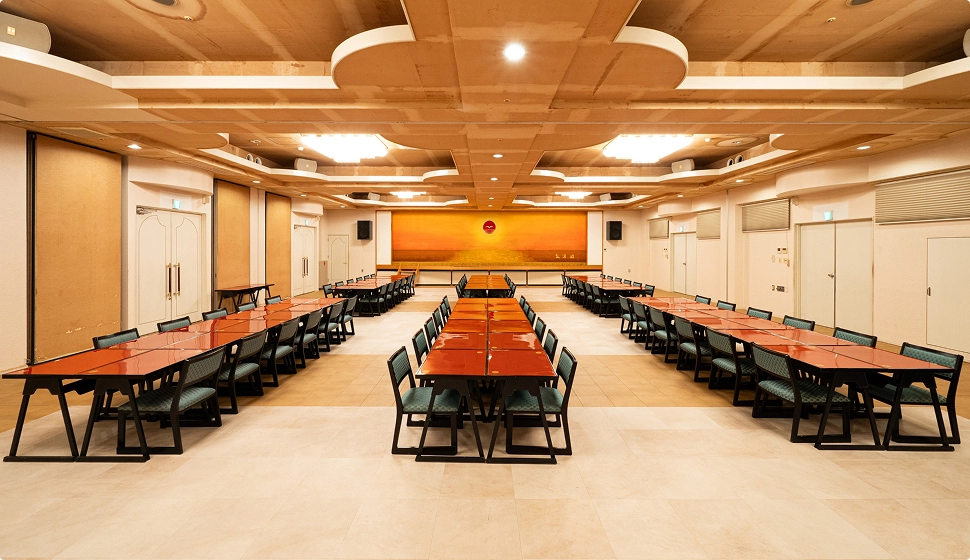 正面に夕日の絵が飾られた広い宴会場に、赤いテーブルと椅子が整然と並べられています。