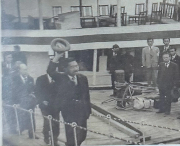 昭和天皇が船のデッキで帽子を高く掲げて挨拶をしています。