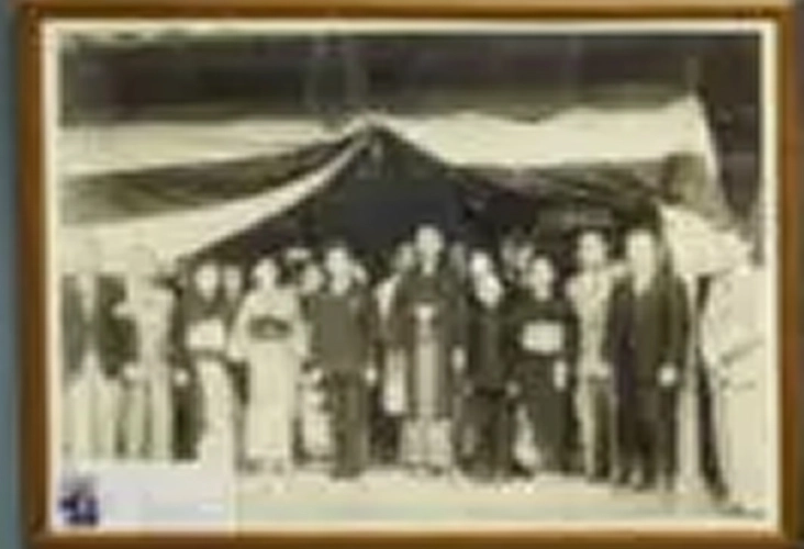 大きな幕の下で、和服や洋服を着た人々が並んで記念撮影をしています。