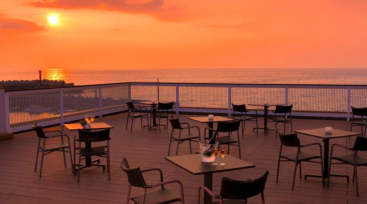 夕日に染まる海辺のテラス席に、飲み物が置かれたテーブルが並んでいます。