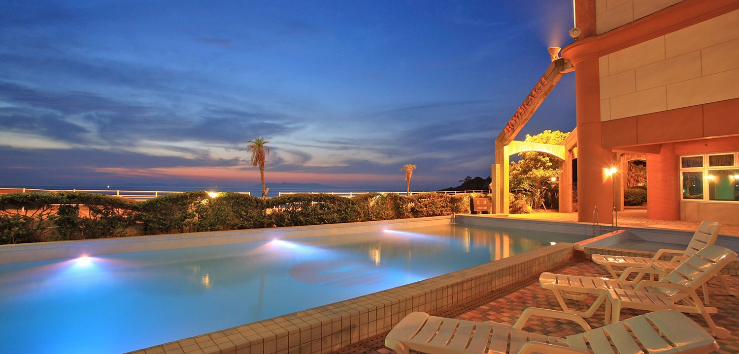 夕暮れ時のリゾートホテルで、ライトアップされたプールサイドにデッキチェアが並んでいます。