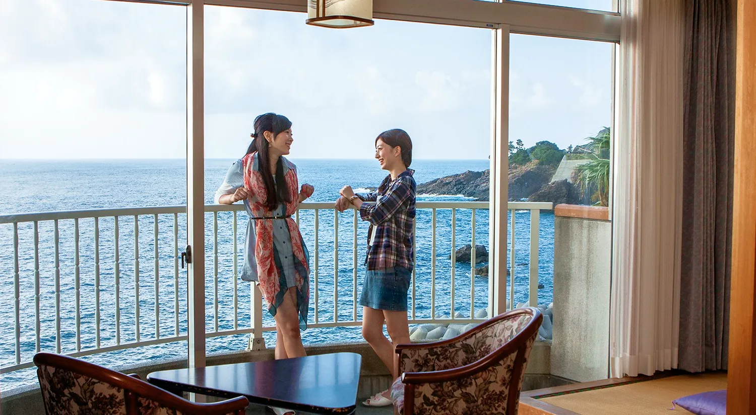 二人の女性がホテルの客室のテラスで海を眺めながら楽しそうに会話をしています。