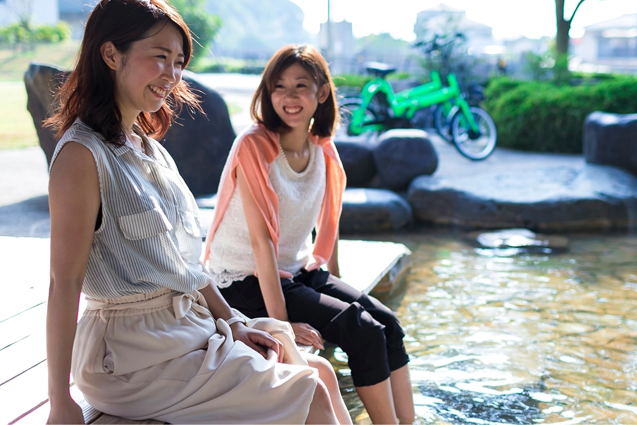 二人の女性が公園の足湯に座り、楽しそうに会話をしています。