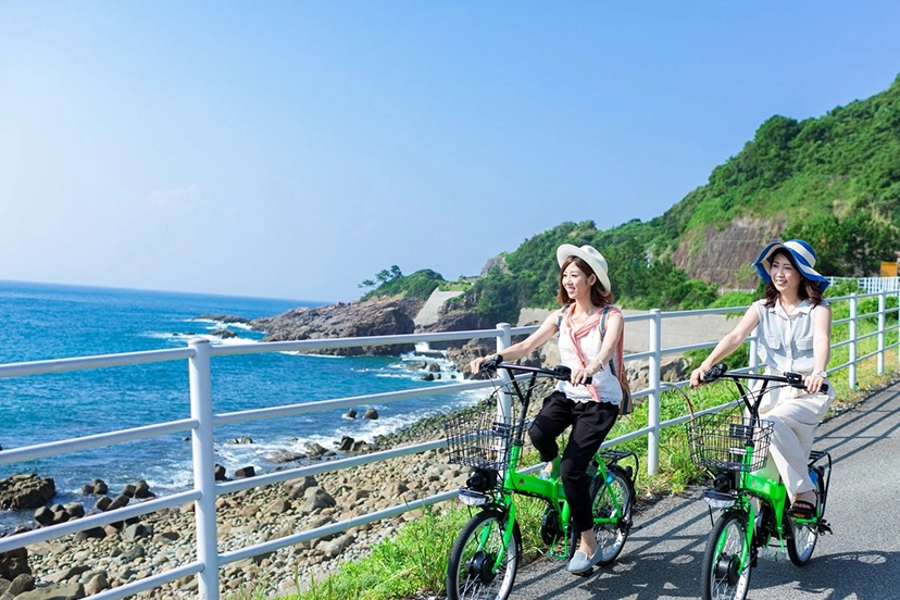 帽子をかぶった二人の女性が、青い海沿いの道を自転車で楽しそうに走っています。