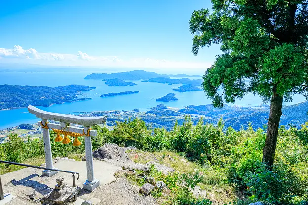 山頂の鳥居越しに、青い海と島々が浮かぶ絶景が広がっています。