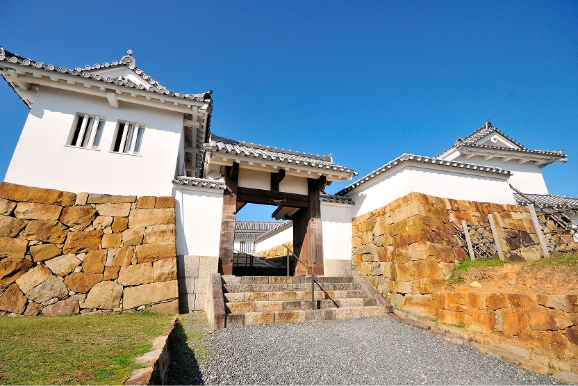 青空の下に白い壁と石垣が美しい、日本の城の門が堂々と建っています。